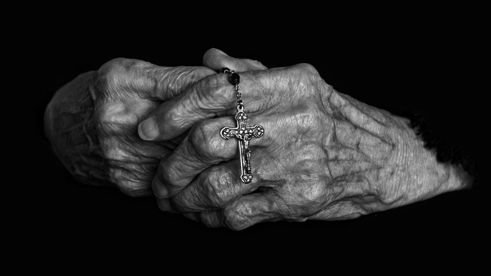 Mãos idosas entrelaçadas segurando um rosário com crucifixo, em preto e branco