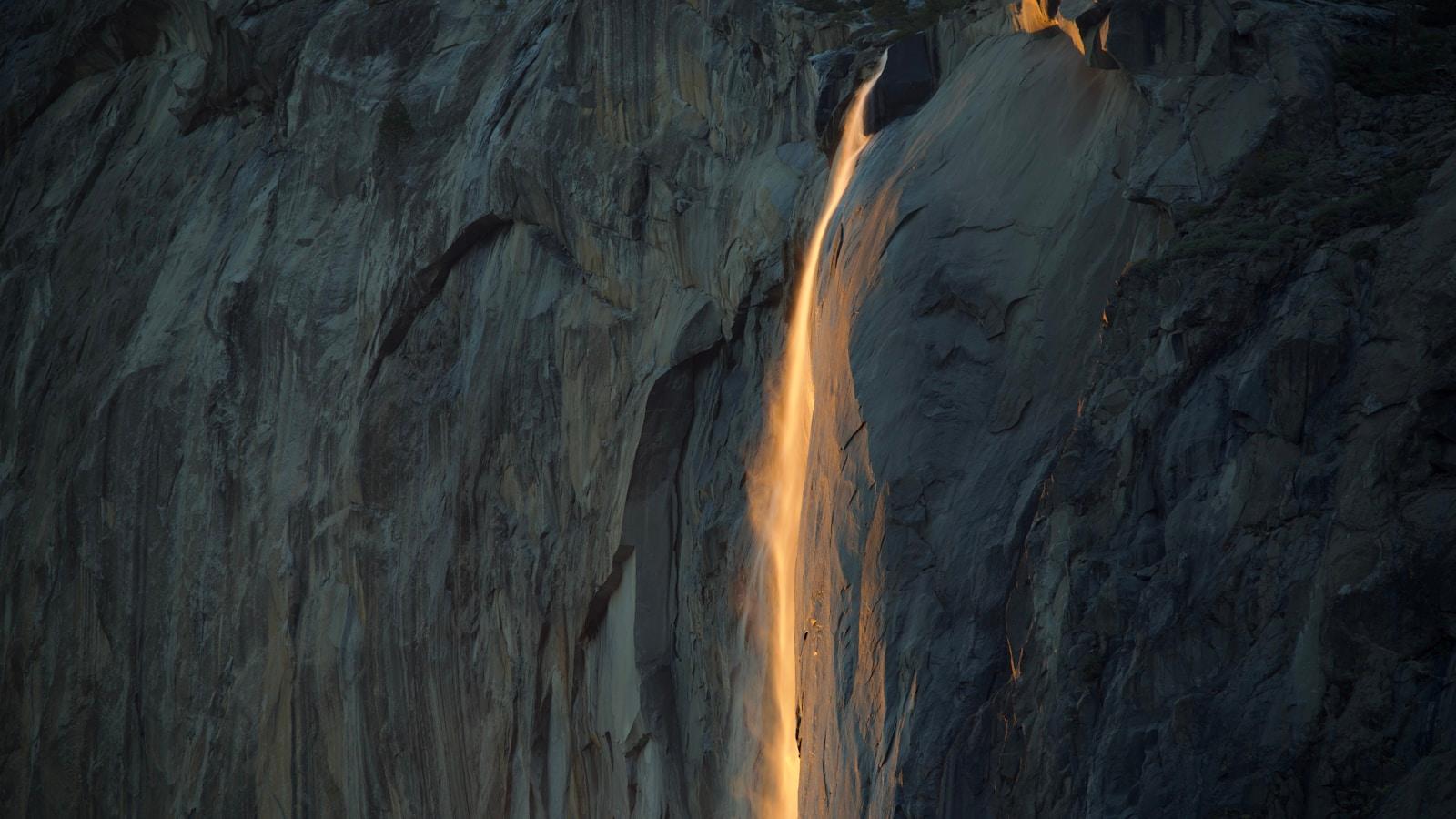 Cachoeira fina descendo por um paredão de rocha, iluminada pela luz alaranjada do sol