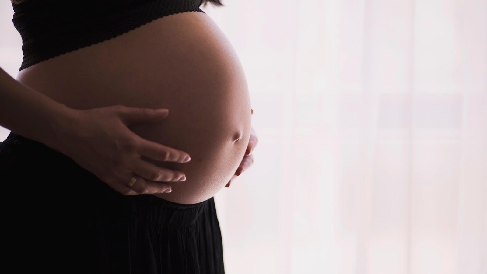 Mulher grávida com as mãos sobre a barriga, diante de uma cortina clara