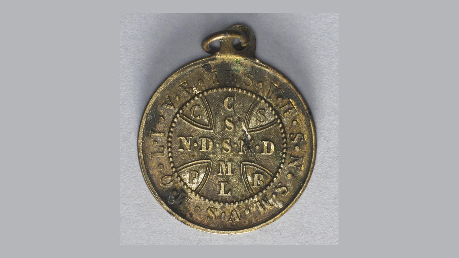 Verso de uma antiga Medalha de São Bento em latão: a cruz com as letras C S S M L e N D S M D, as iniciais em volta e IHS no alto