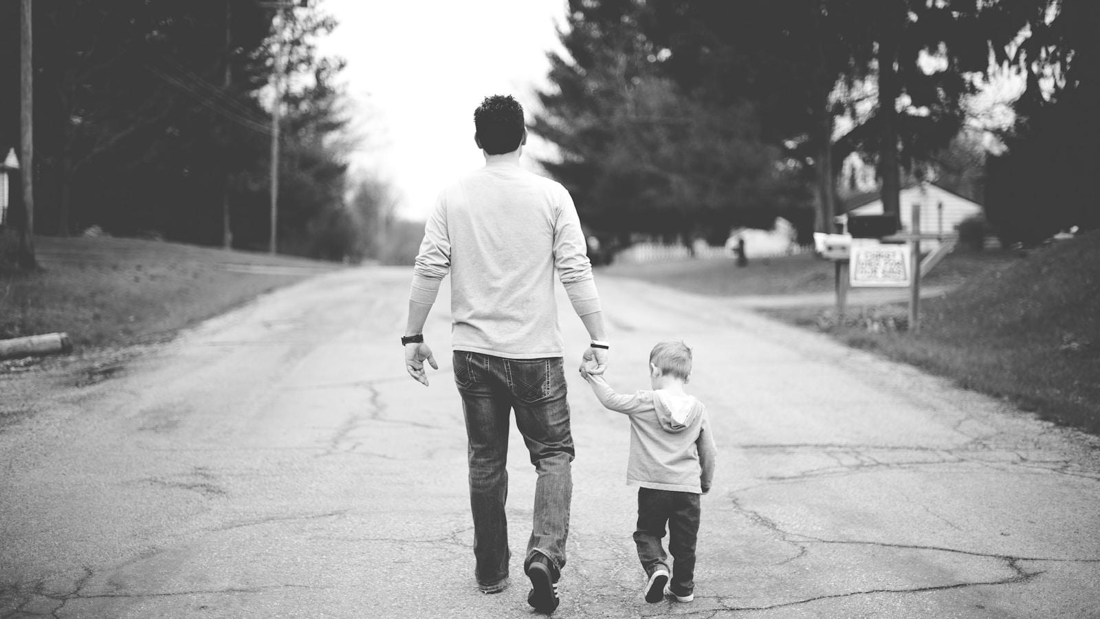 Pai e filho pequeno caminhando de mãos dadas por uma rua, em preto e branco