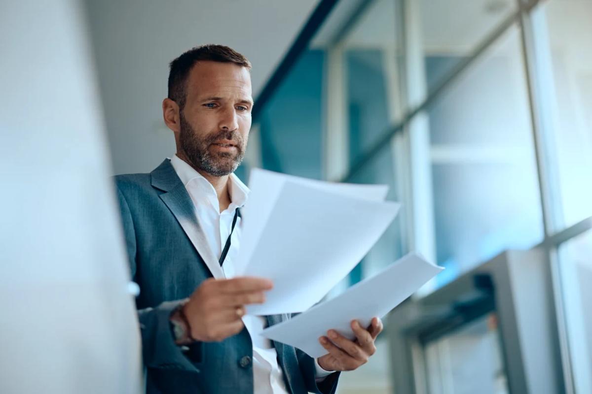 A man in a suit standing in a bright glass-walled office, reading through loose printed pages