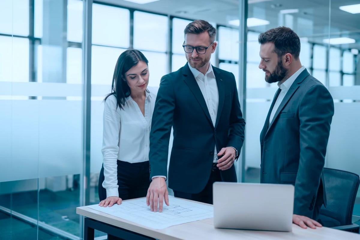 Three colleagues in business dress around a table with a laptop and a printed plan in a glass meeting room