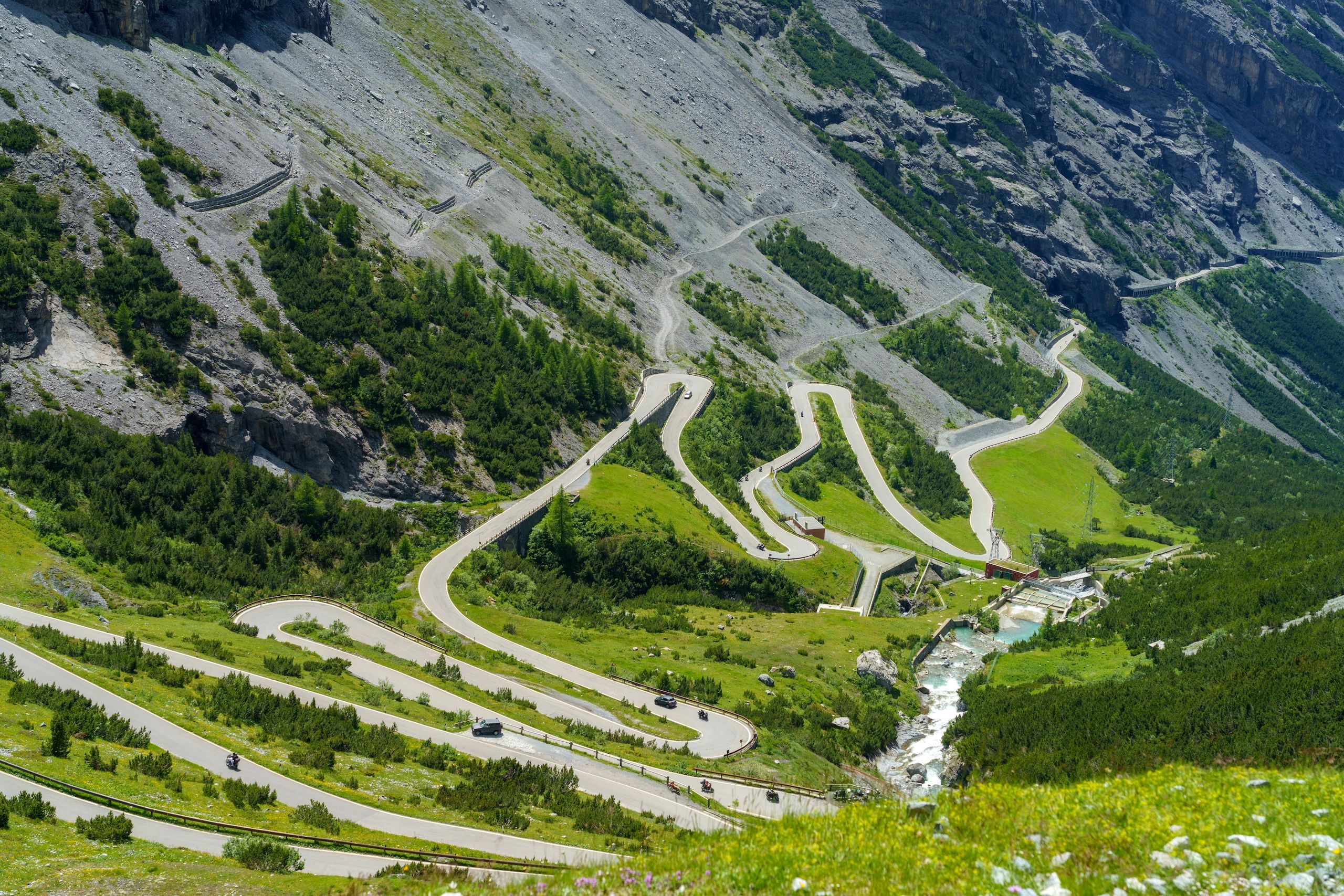 Passo Stelvio på vei til Bormio. Svingete fjellveier med grønne daler og bratte klippevegger. En elv renner gjennom landskapet, og små biler kan sees på veiene som snor seg gjennom naturen.