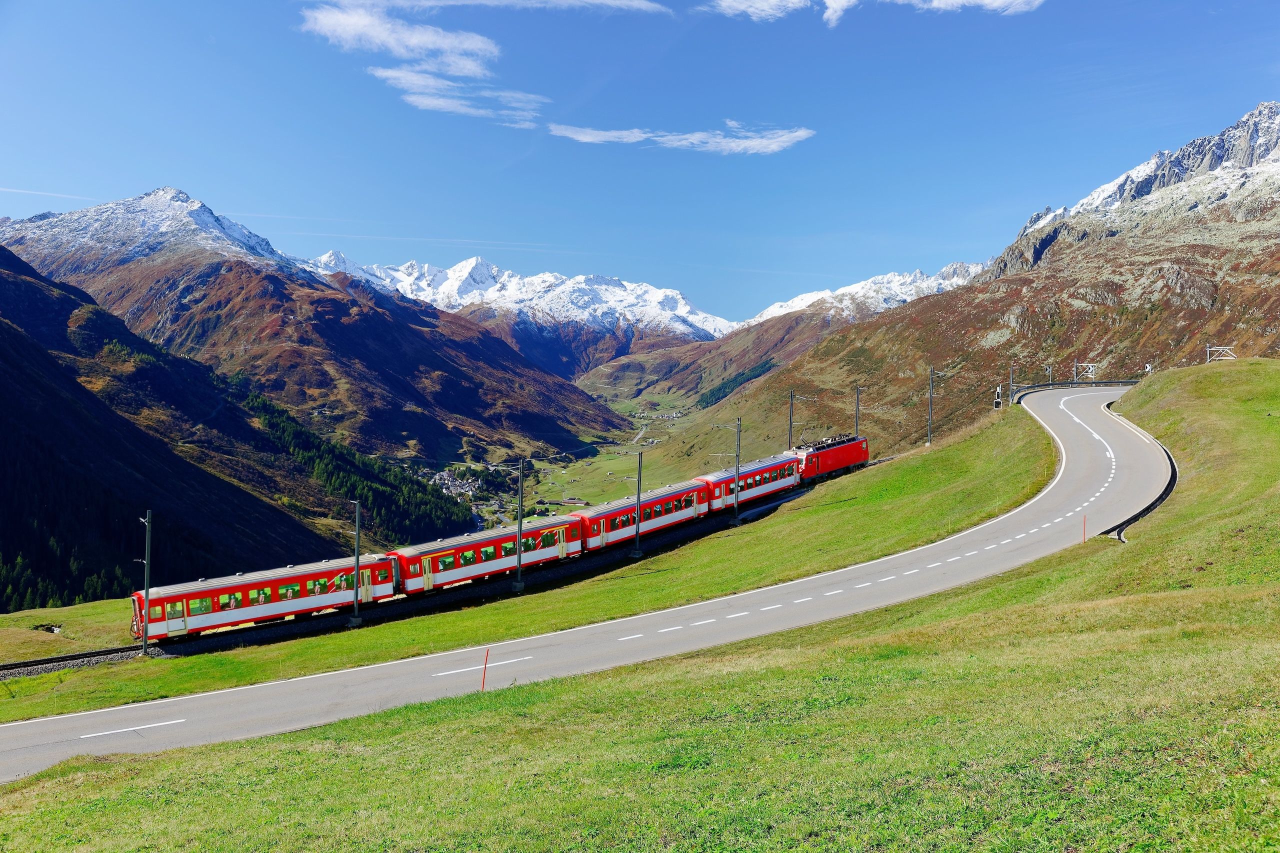 Landevei på vei til Oberalppass, nydelig utsikt mot Andermatt og snøtopper på fjellene blant grønne enger. I tillegg er det røde, velkente toget Galcier express også i bildet.