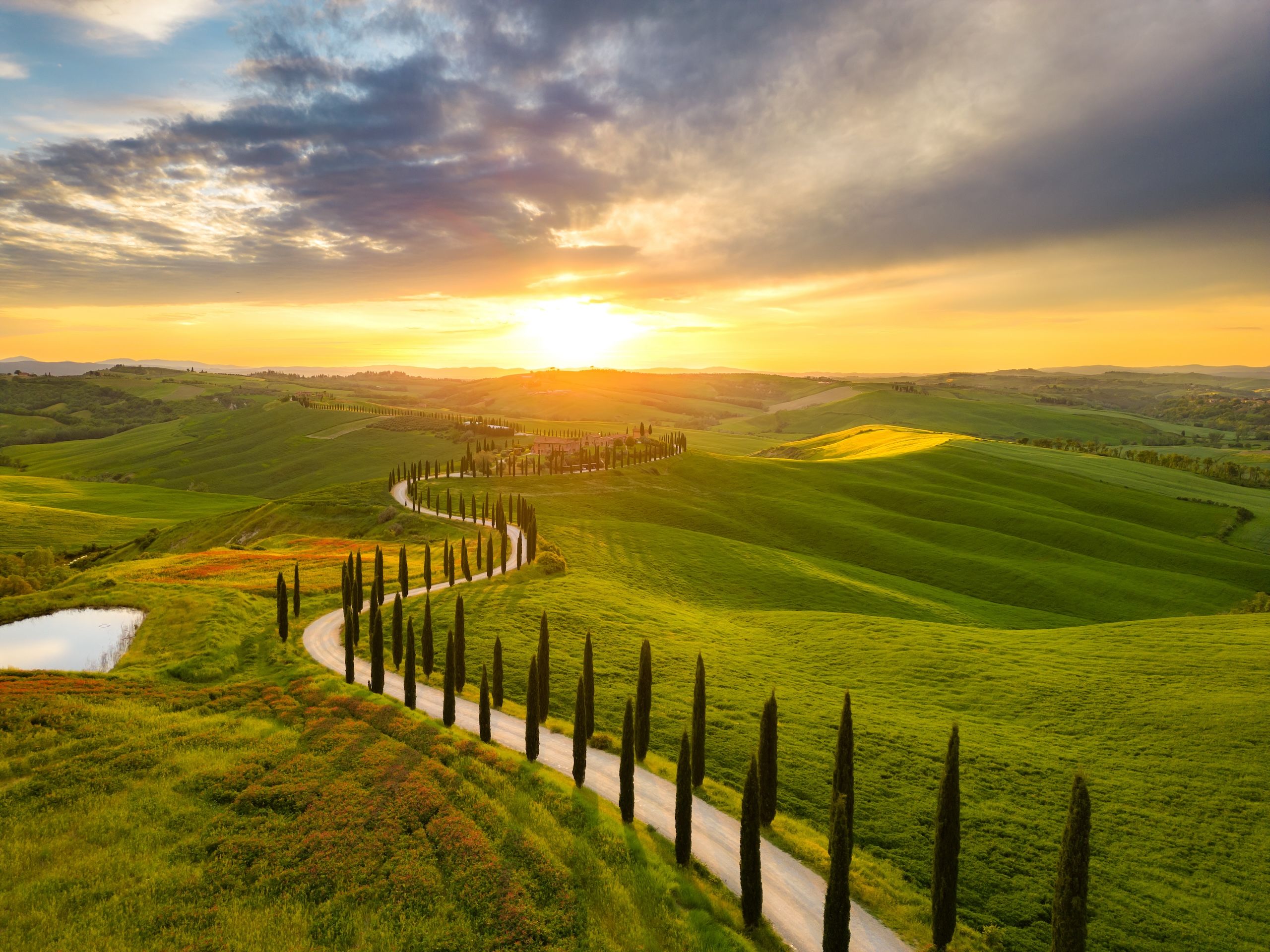 Solnedgang over de bølgende åsene i Toscana, Italia, med cypresser som linjer en grusvei gjennom landskapet.