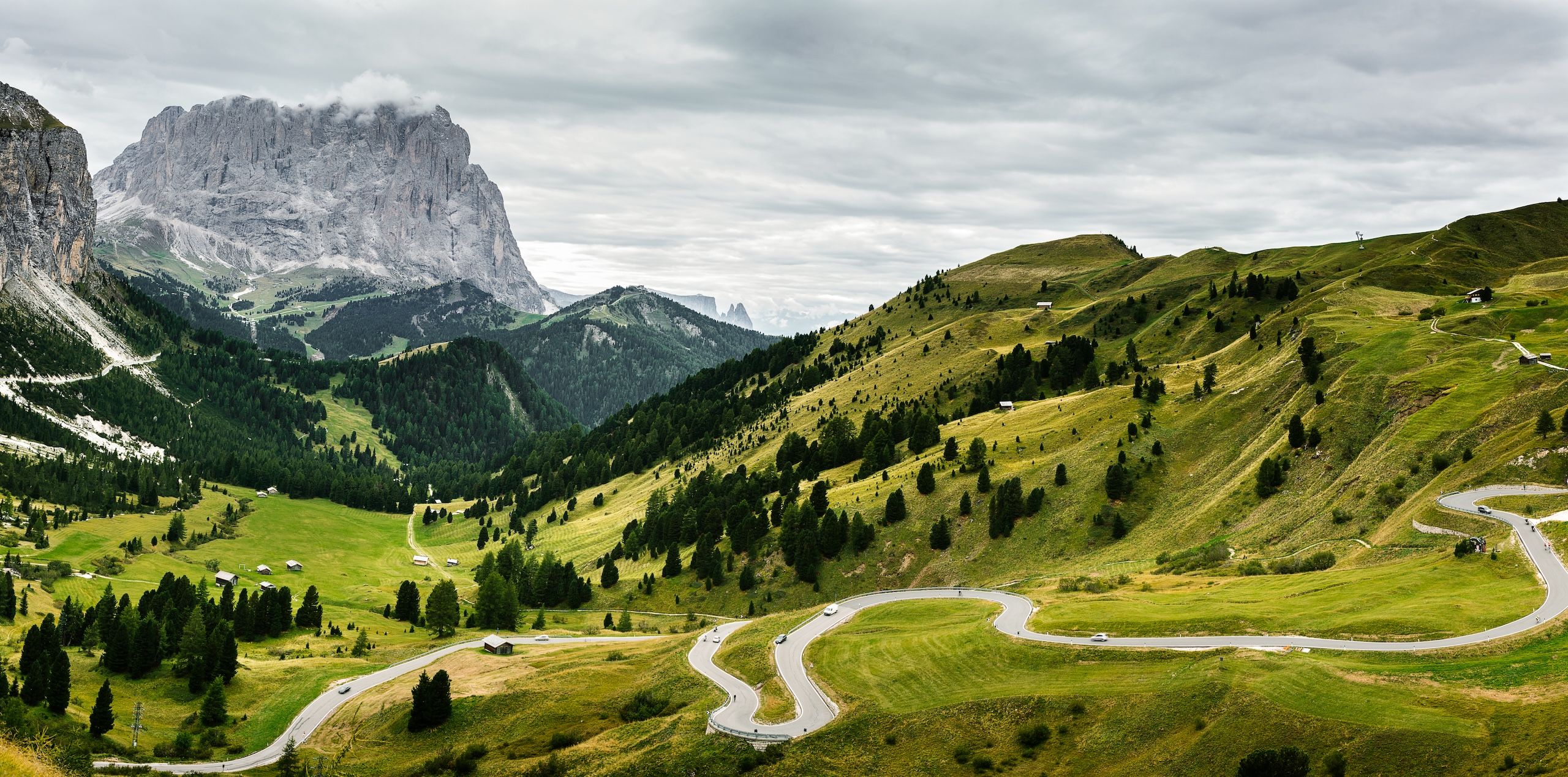 Svingete veier som snor seg gjennom de grønne Dolomittene i Italia med dramatiske fjell i bakgrunnen under en overskyet himmel.
