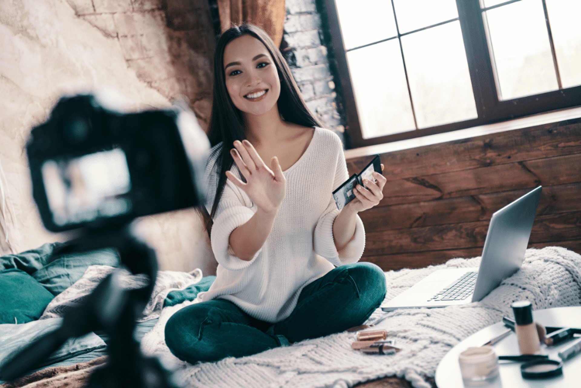 A woman is seated on her bed and is looking at her video camera. Her laptop is open next to her.