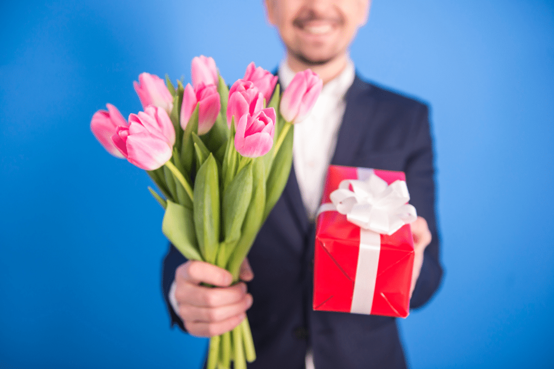 A gentleman in a suit extends flowers and a present to the viewer