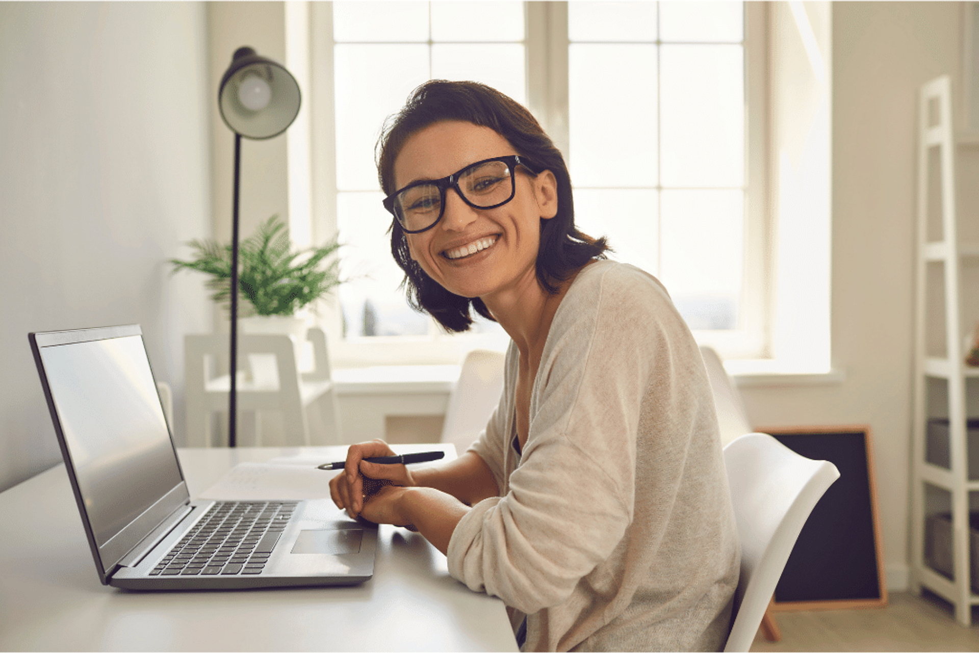 A woman is smiling at the camera while sitting at her desk with a laptop in front of her