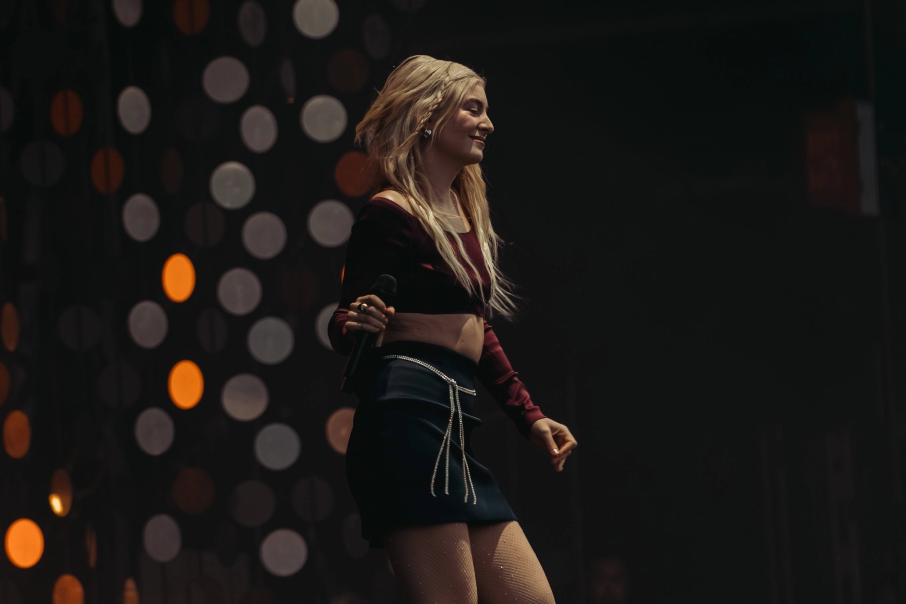 Lorde smiling on stage in a cropped red top and dark mini skirt, warm bokeh lights behind her