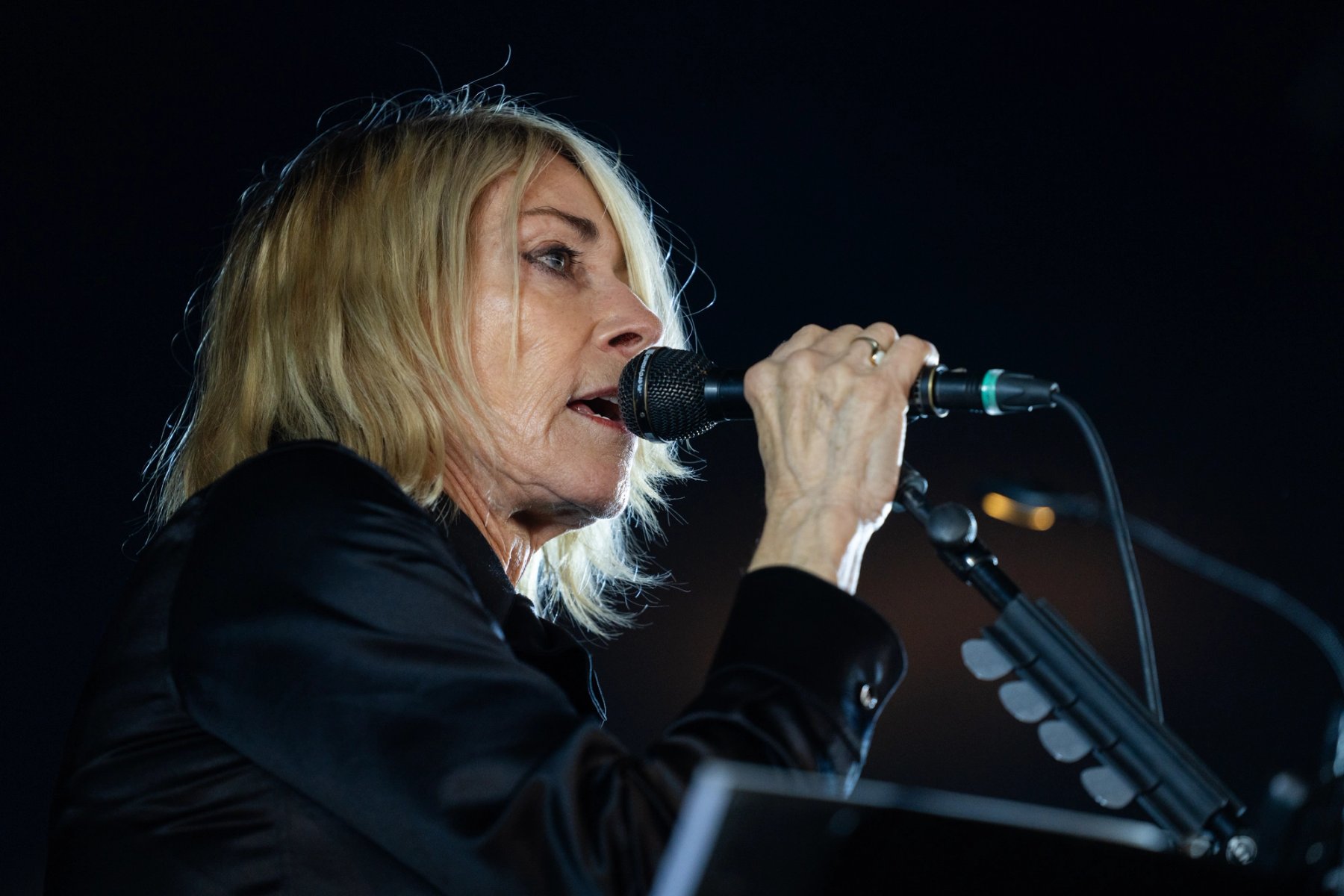 Kim Gordon singing into a microphone in profile, in a black jacket against a dark background