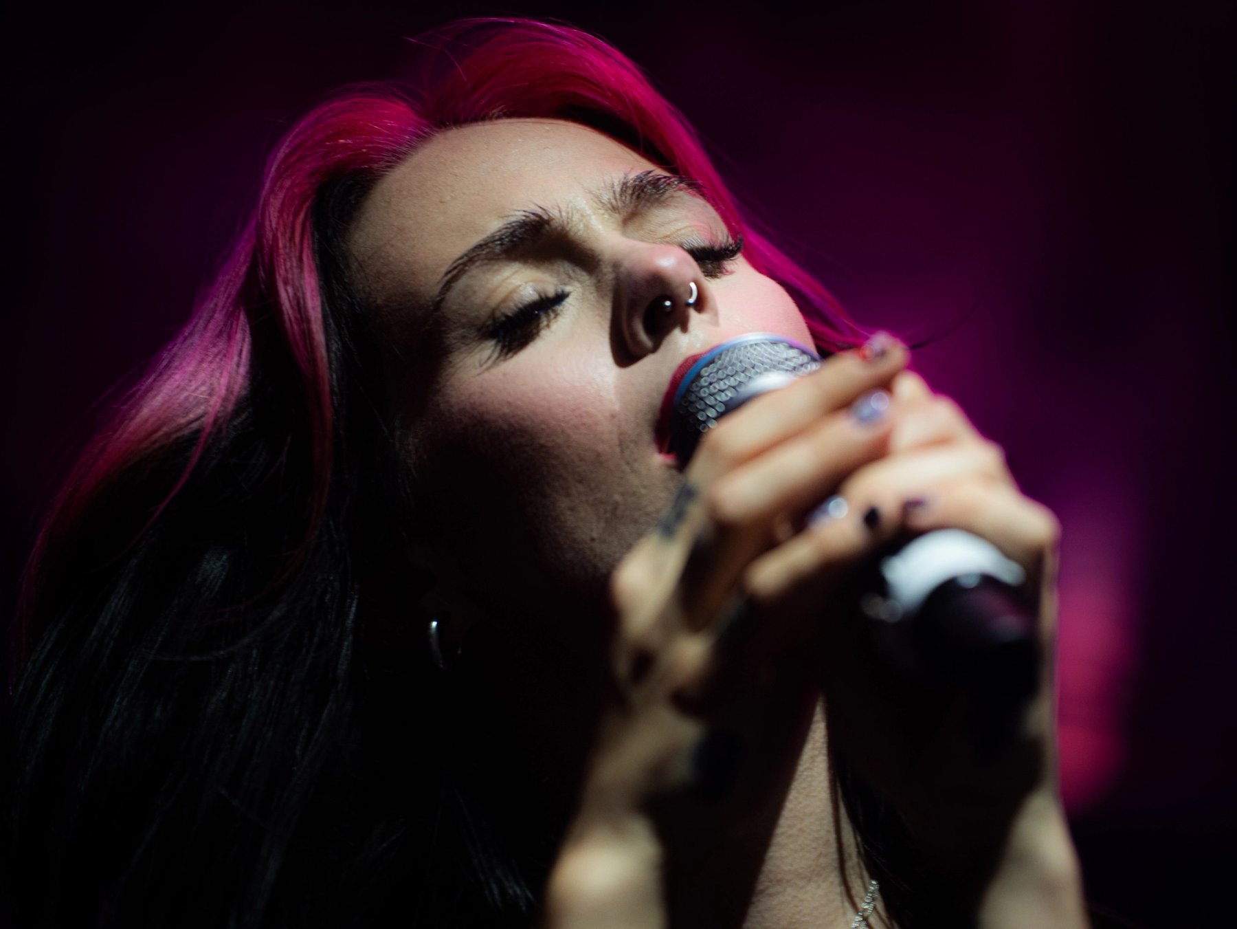 Close-up of Girli singing with her eyes closed under magenta light