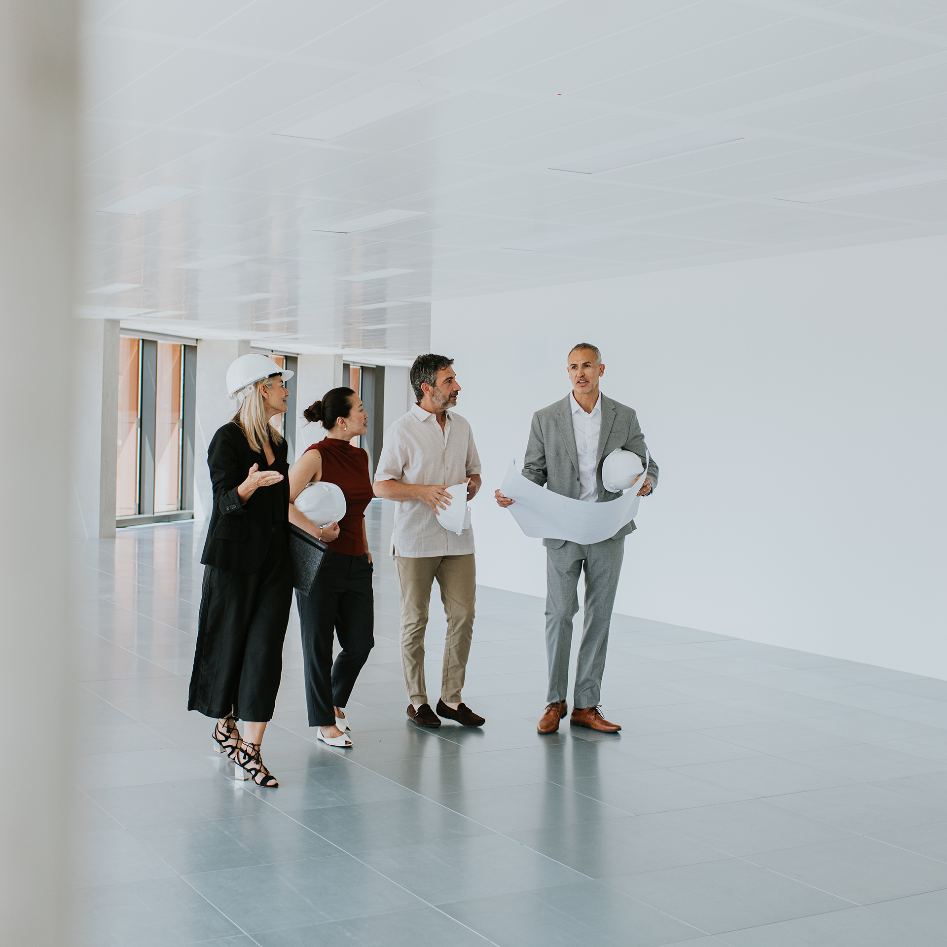 A project team walks through a modern commercial office building during a construction site walkthrough. An architect carrying rolled construction plans and a white safety helmet leads the discussion with colleagues, including a construction manager and clients holding safety helmets, as they inspect the unfinished interior and discuss project progress.