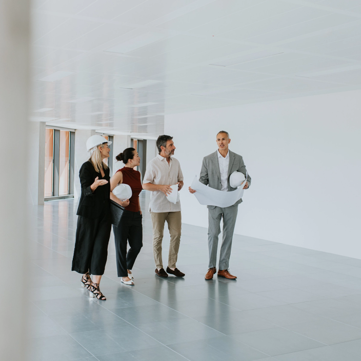 A project team walks through a modern commercial office building during a construction site walkthrough. An architect carrying rolled construction plans and a white safety helmet leads the discussion with colleagues, including a construction manager and clients holding safety helmets, as they inspect the unfinished interior and discuss project progress.