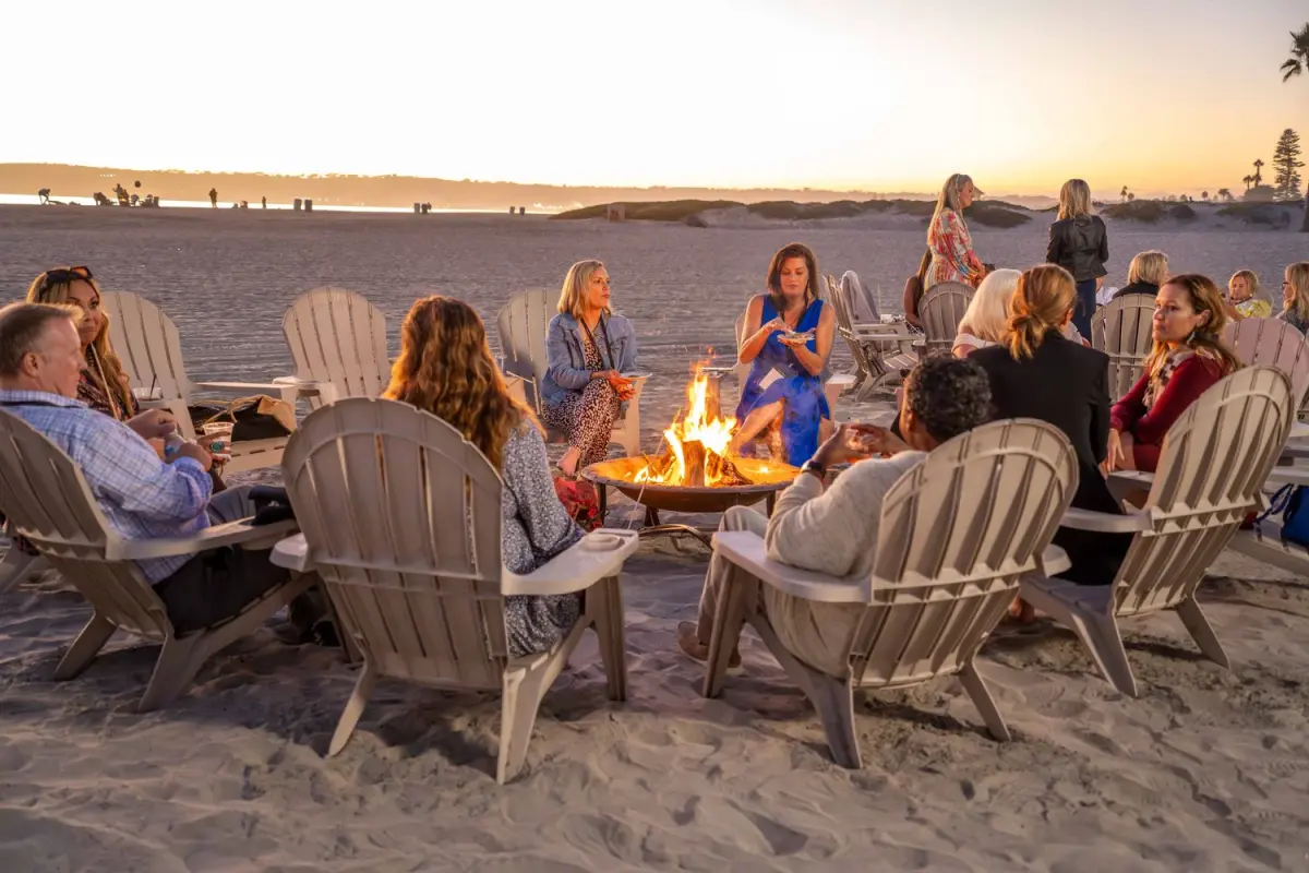 Attendees gathered around a beach fire at dusk, enjoying drinks and conversation during KW Luxury reception.