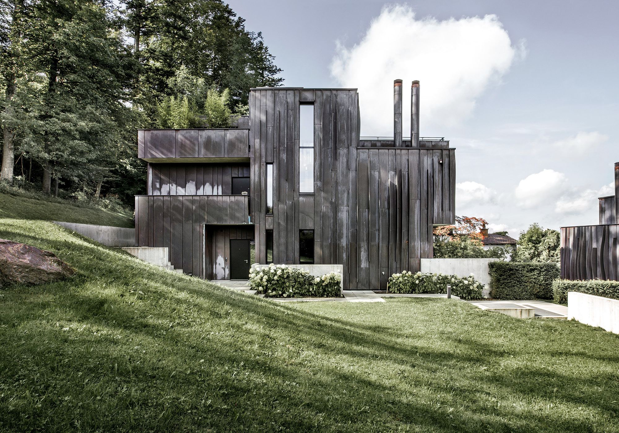Dark volumes with chimneys on the slope – Apartment building in Uitikon