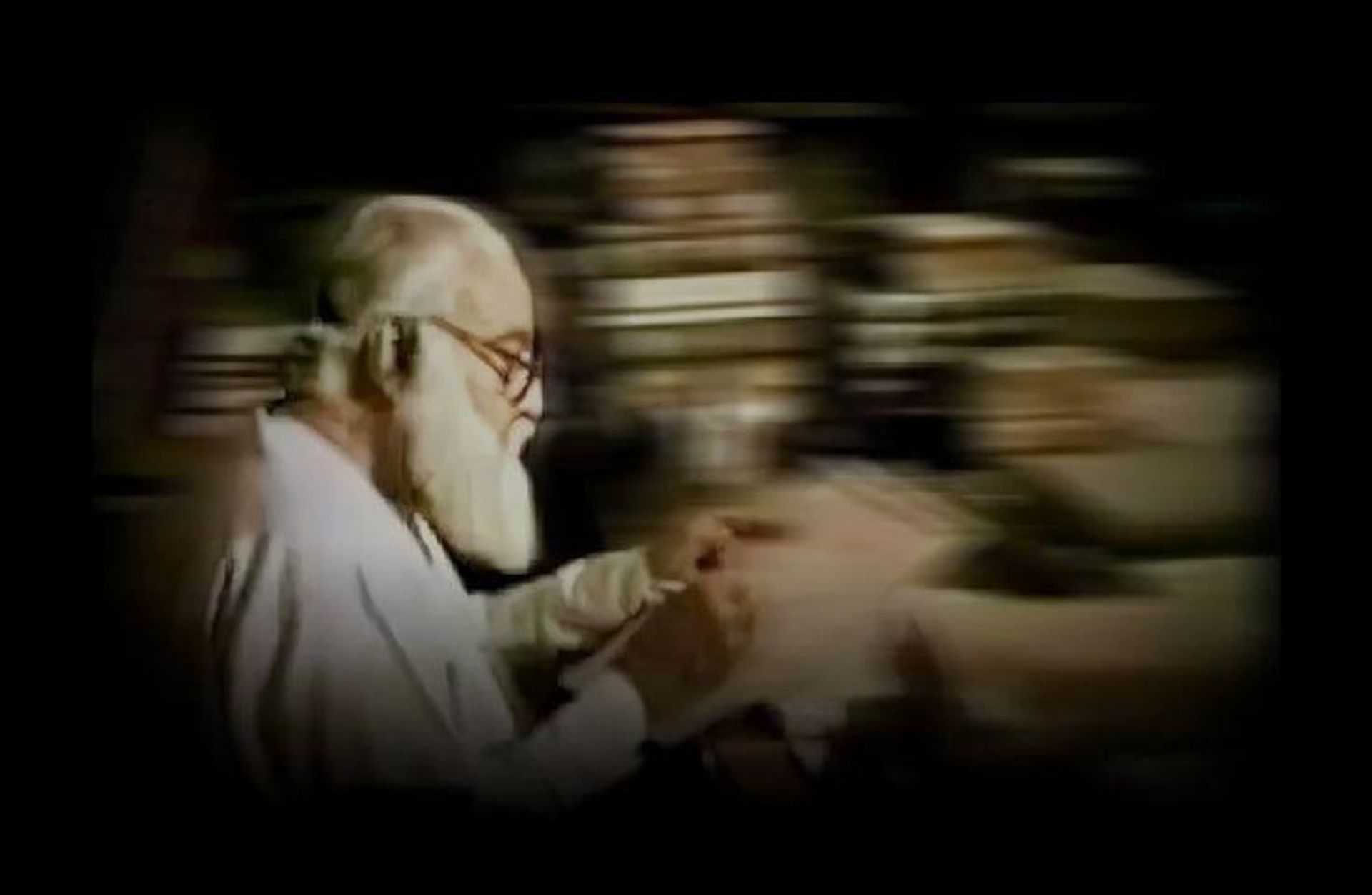 Elderly man with white beard and glasses reads a document against a motion-blurred background.