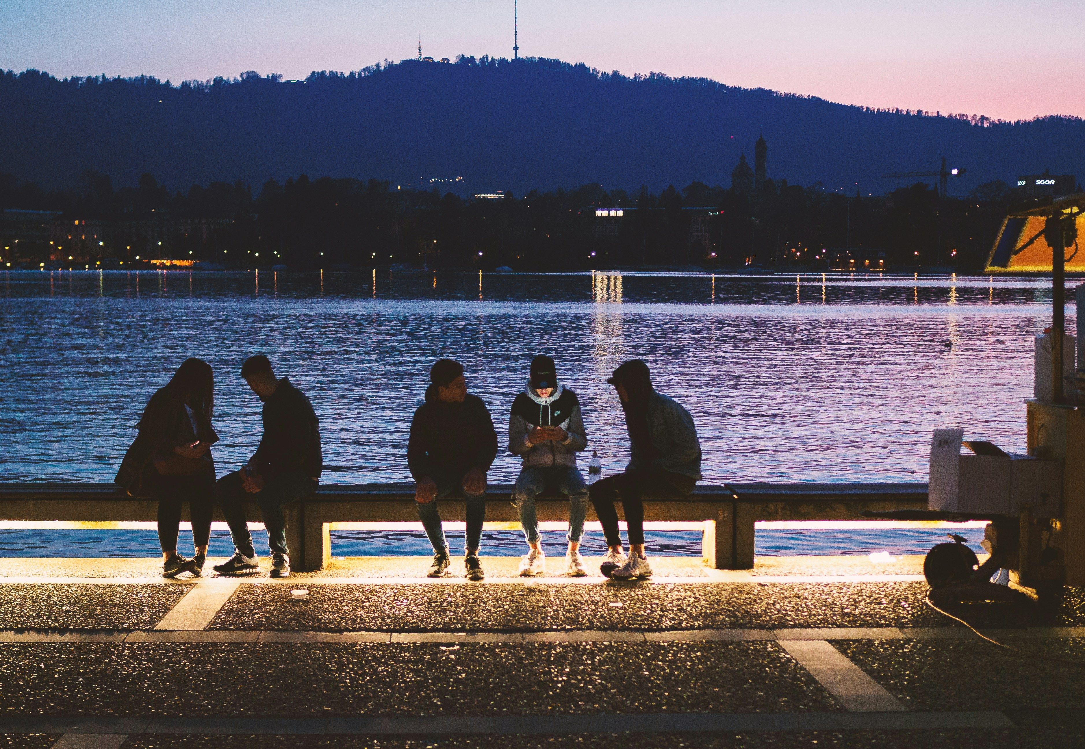 Four silhouetted people sit by a lake at dusk, with city lights and mountains in the distance.