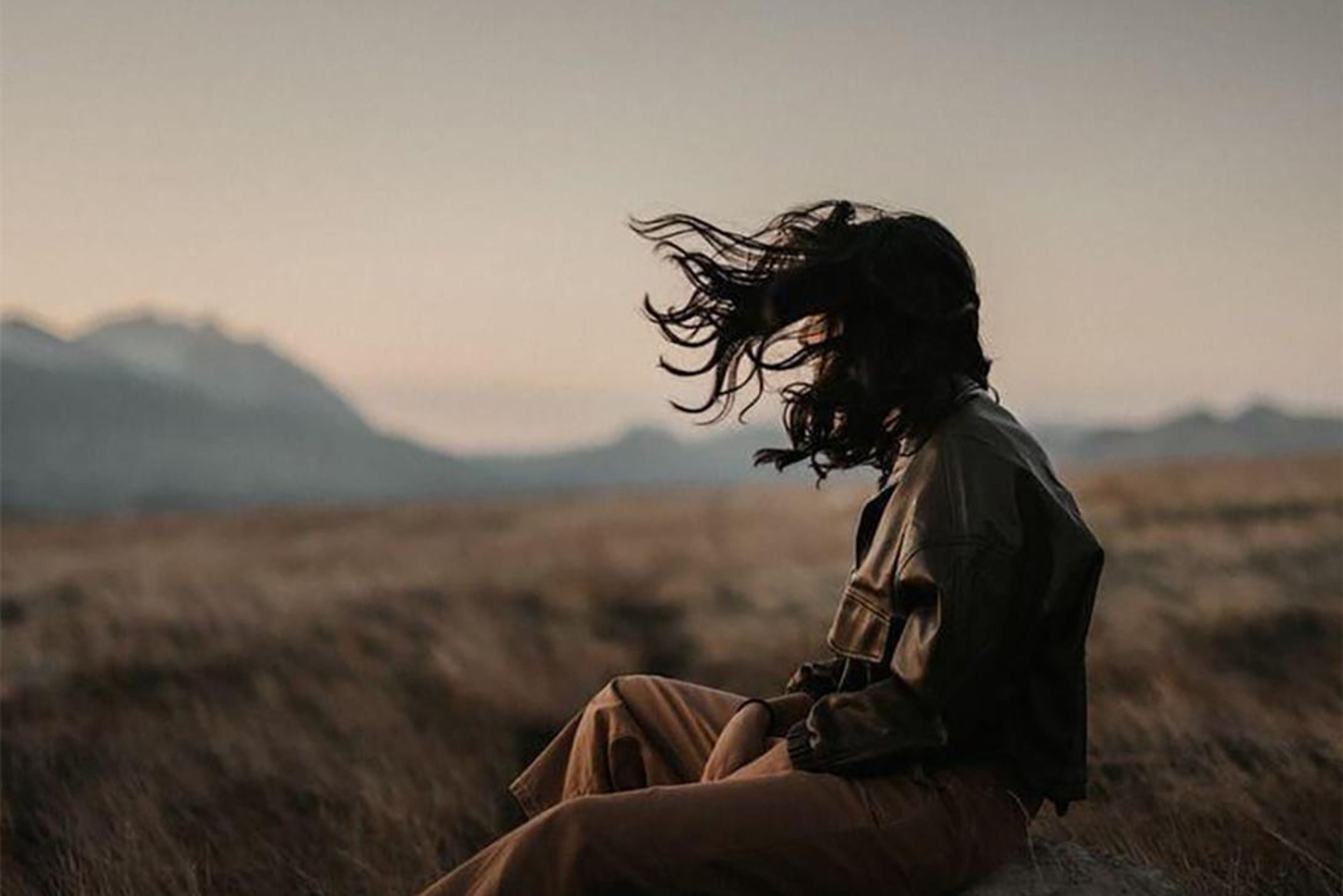 Profile of a person with windblown hair sitting in a dry field, mountains in the distance.