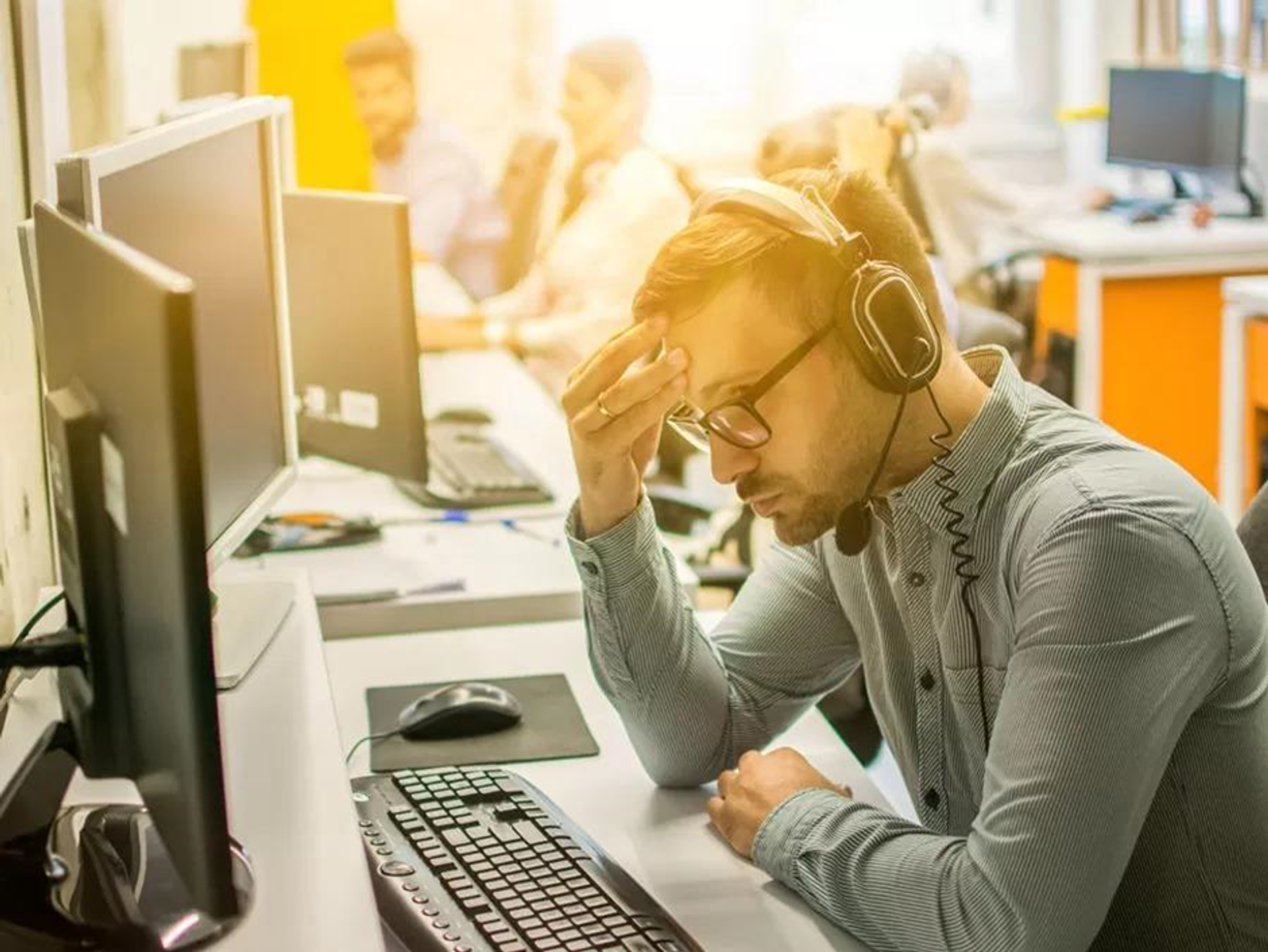 A man in a headset looks stressed while sitting at a computer in an office.