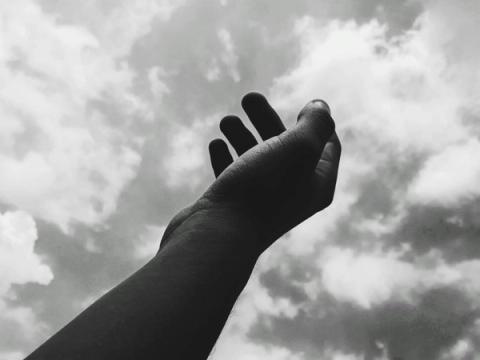 Black and white image of a hand reaching up towards a cloudy sky.