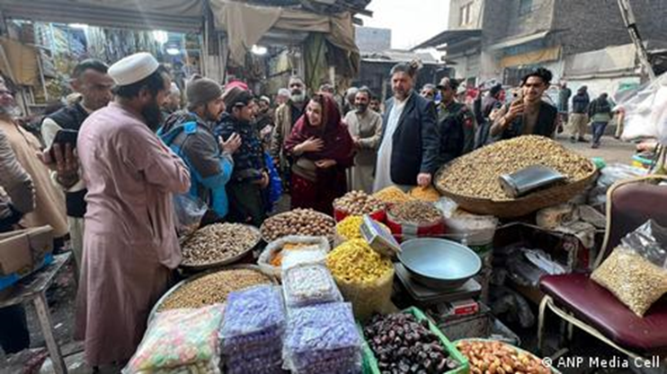 A bustling outdoor market with people browsing stalls piled high with nuts, dried fruits, and snacks. A woman in a red outfit and a man in a suit are visible among the crowd.
