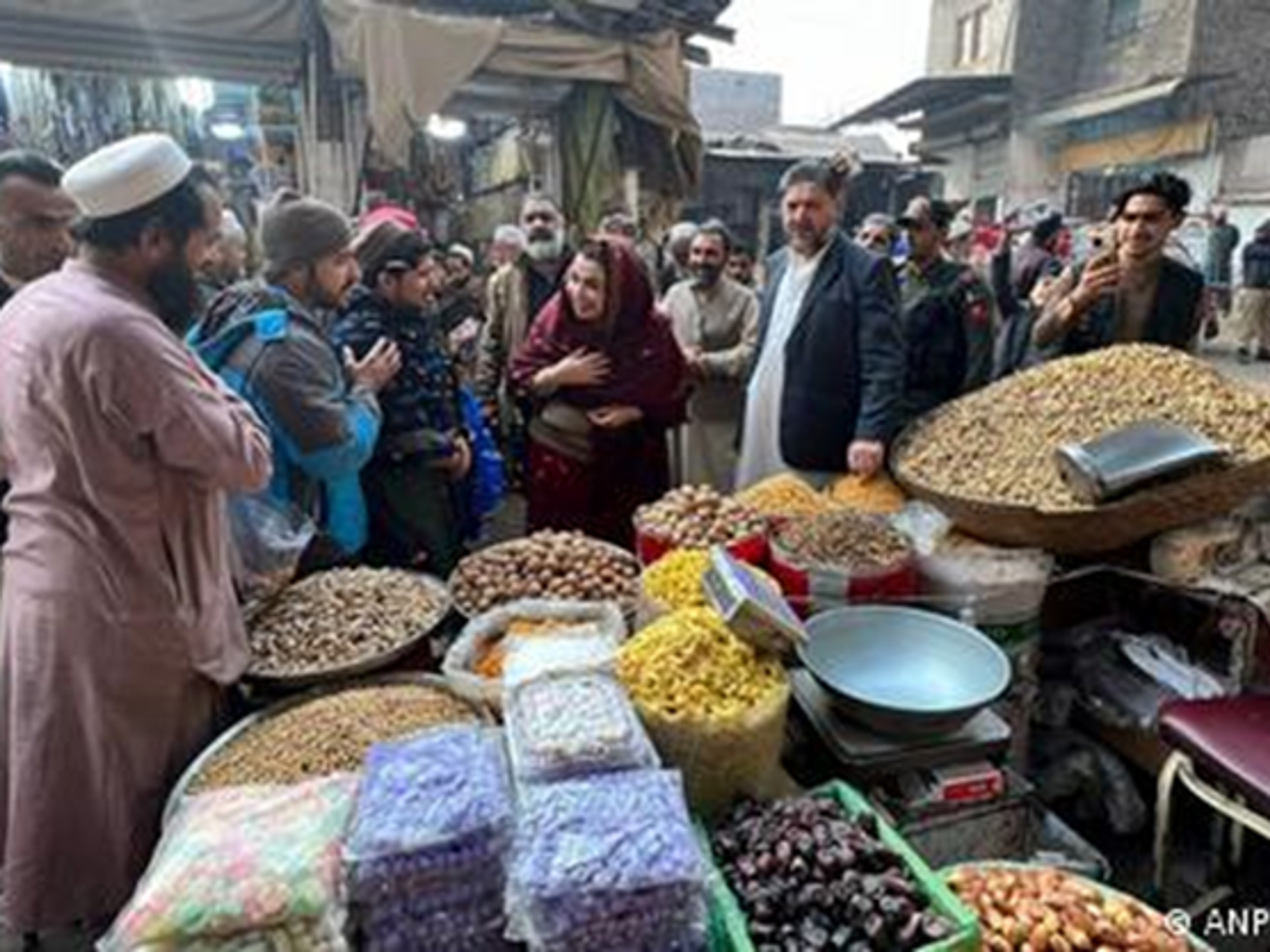 A bustling outdoor market with people browsing stalls piled high with nuts, dried fruits, and snacks. A woman in a red outfit and a man in a suit are visible among the crowd.