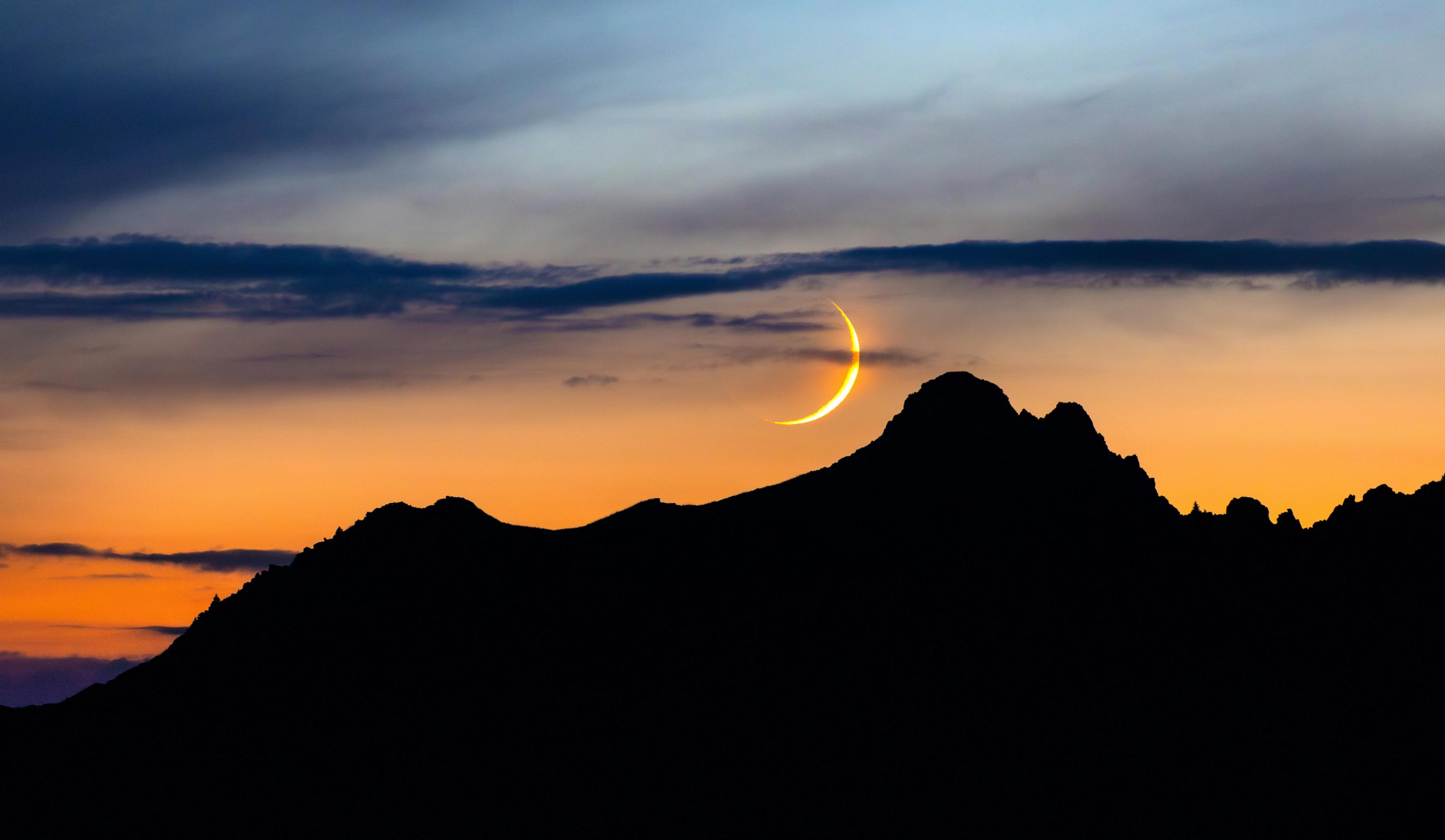 Thin crescent moon over silhouetted mountains at sunset.