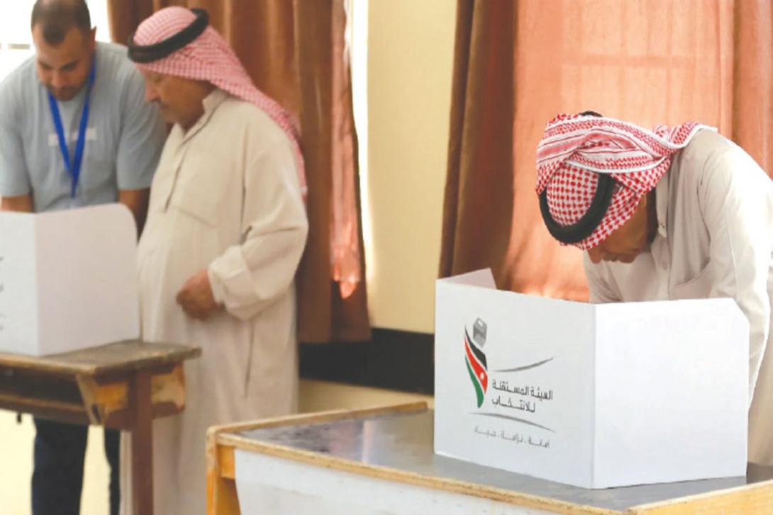 Two men in traditional Middle Eastern dress stand at individual voting booths, one bending to cast a ballot, with an election official nearby.