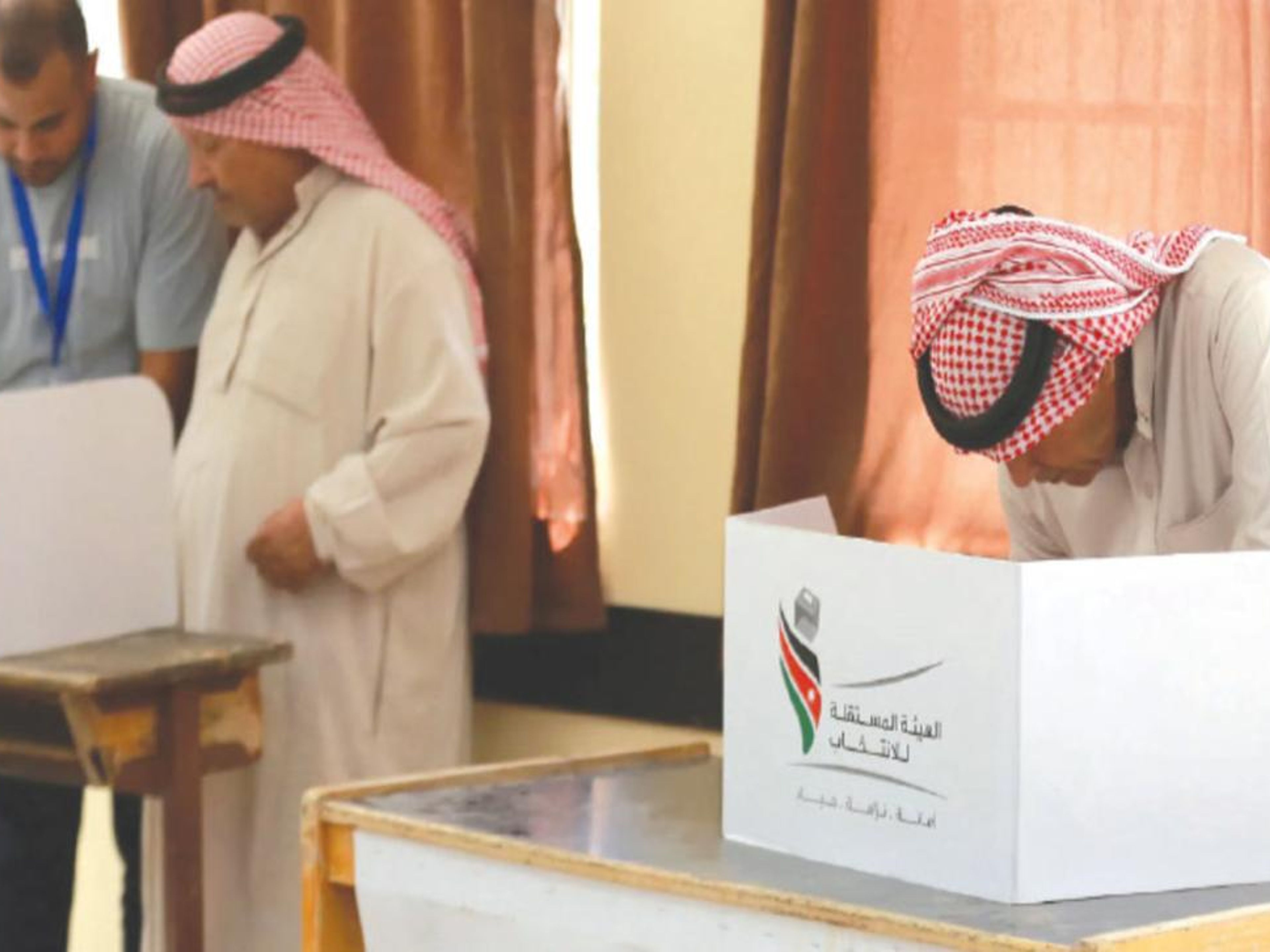 Two men in traditional Middle Eastern dress stand at individual voting booths, one bending to cast a ballot, with an election official nearby.