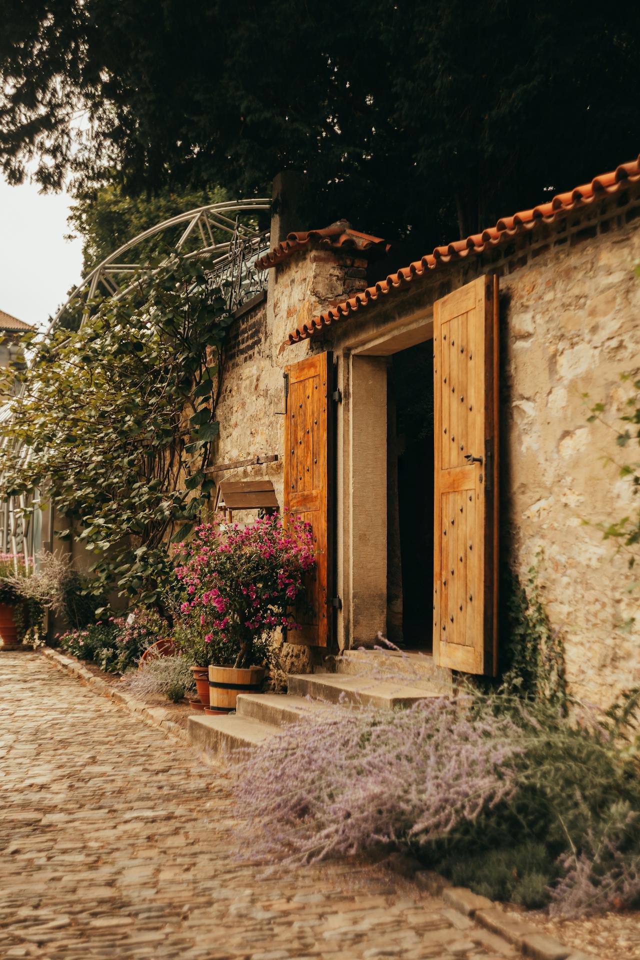 Rustic stone building with open wooden doors, surrounded by pink and purple flowers and a cobblestone path.
