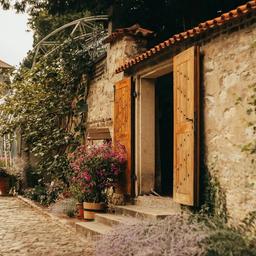 A rustic stone building with an open wooden door, blooming pink and purple flowers, and a cobblestone path.