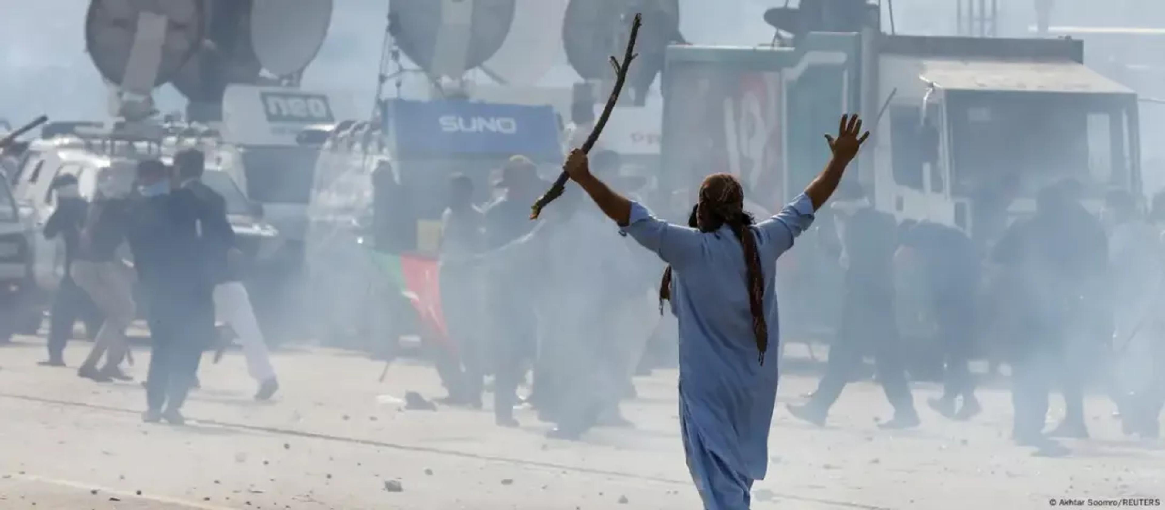 A man in traditional dress holds a stick and raises an arm in a smoky street, with news vans behind.
