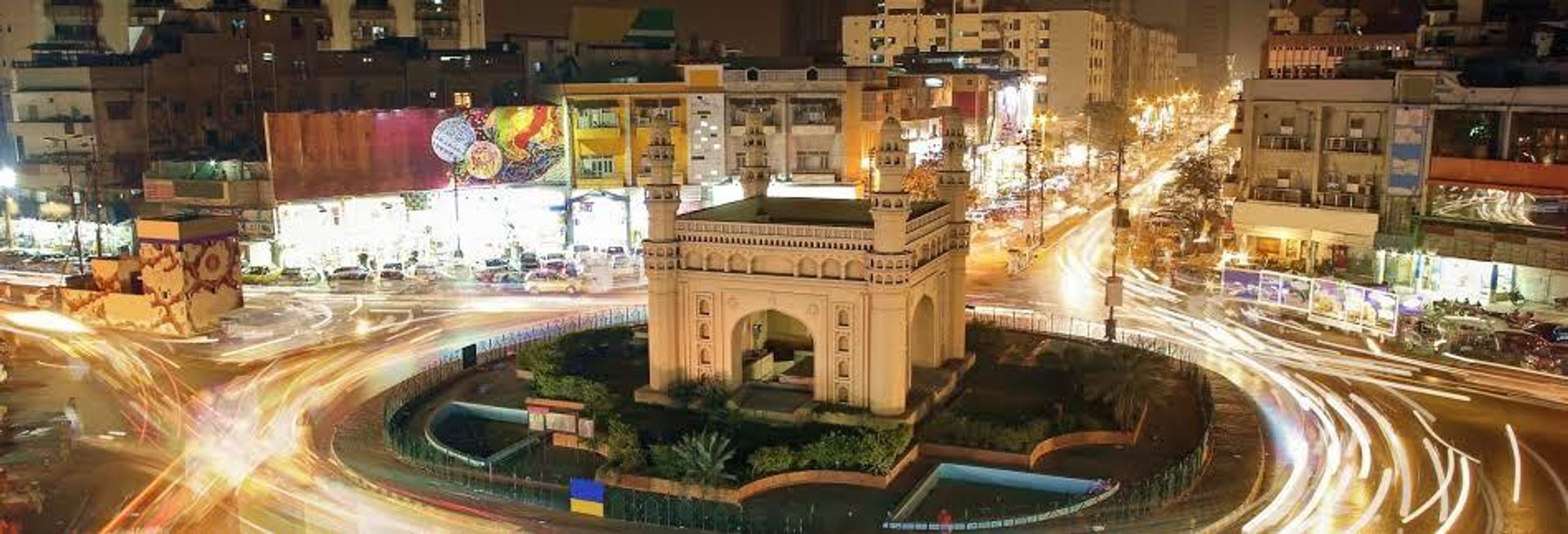 Night long exposure of a Charminar-style monument in a busy city roundabout with vehicle light trails.