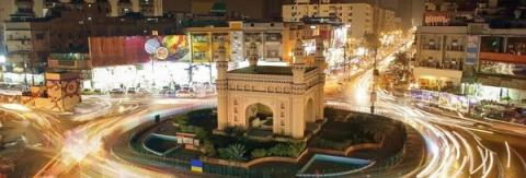 Night long exposure of a Charminar-style monument in a busy city roundabout with vehicle light trails.