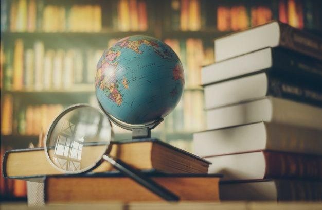 A globe, magnifying glass, and stacks of books on a desk, with bookshelves in the background.