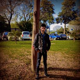 A young man in a dark jacket stands next to a tree in a sunny park with cars in the background.