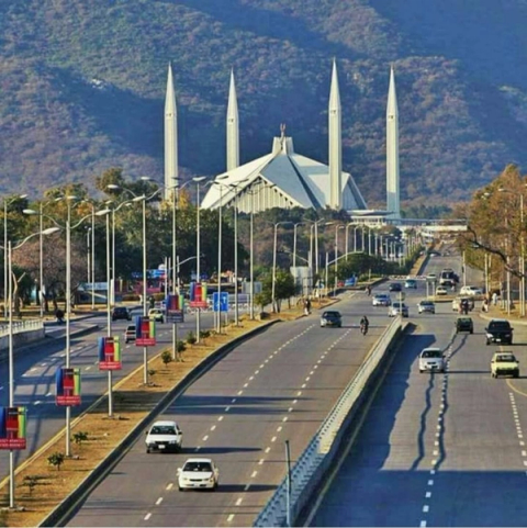 A wide road with cars leads to the modern white Faisal Mosque with four tall minarets, set against forested hills.