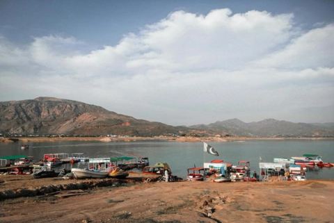 Boats moored at a lakeshore with a Pakistani flag and mountains in the background.