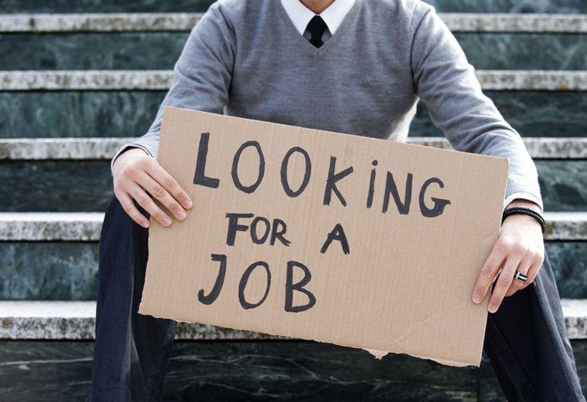 Person sitting on steps, holding a cardboard sign that reads "LOOKING FOR A JOB."
