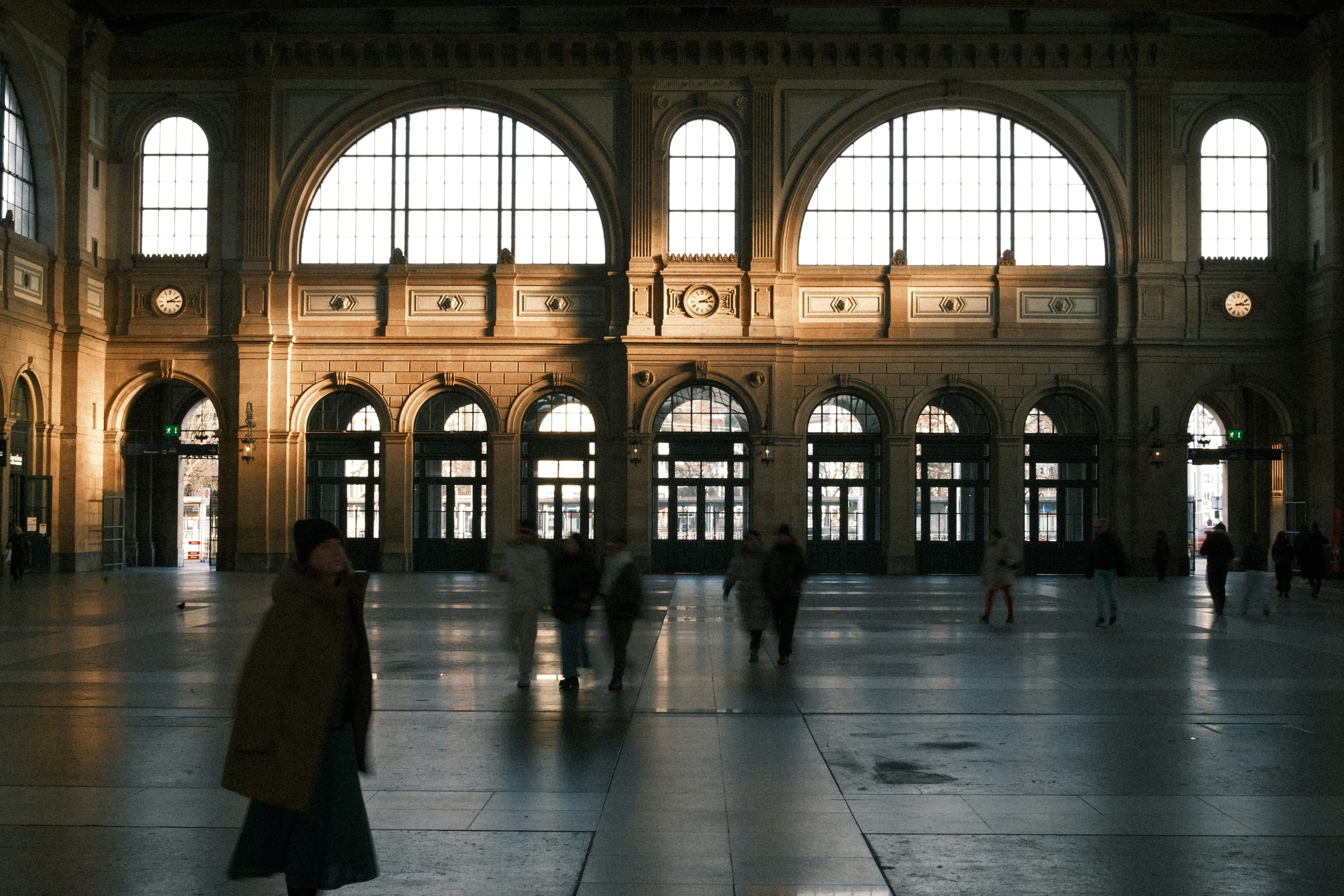 Grand, ornate train station interior with large arched windows, sunlight, and people in motion.