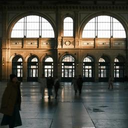 Grand, ornate train station interior with large arched windows, sunlight, and people in motion.