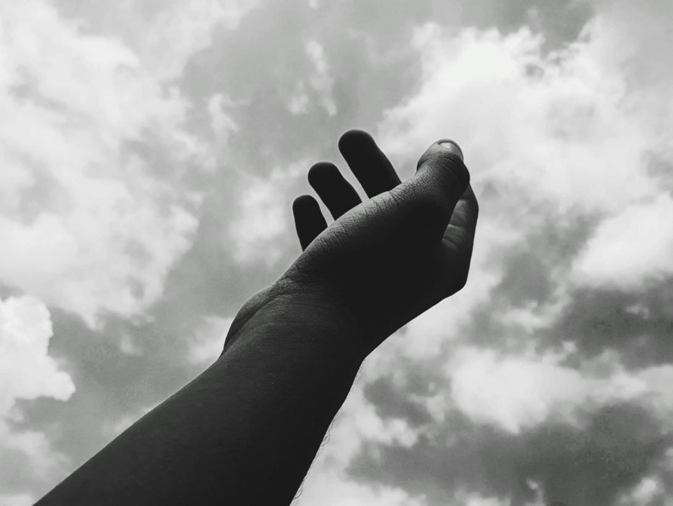 Black and white photo of an arm and open hand reaching up against a cloudy sky.