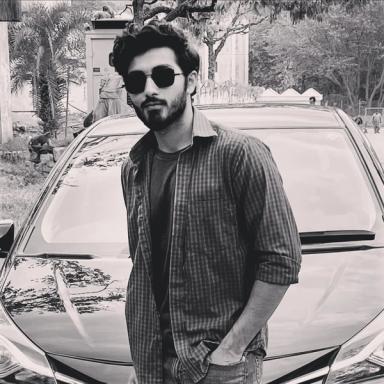 Black and white image of a young man with a beard and sunglasses, wearing a plaid shirt, standing in front of a car.