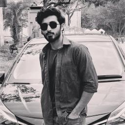 Black and white image of a young man with a beard and sunglasses, wearing a plaid shirt, standing in front of a car.