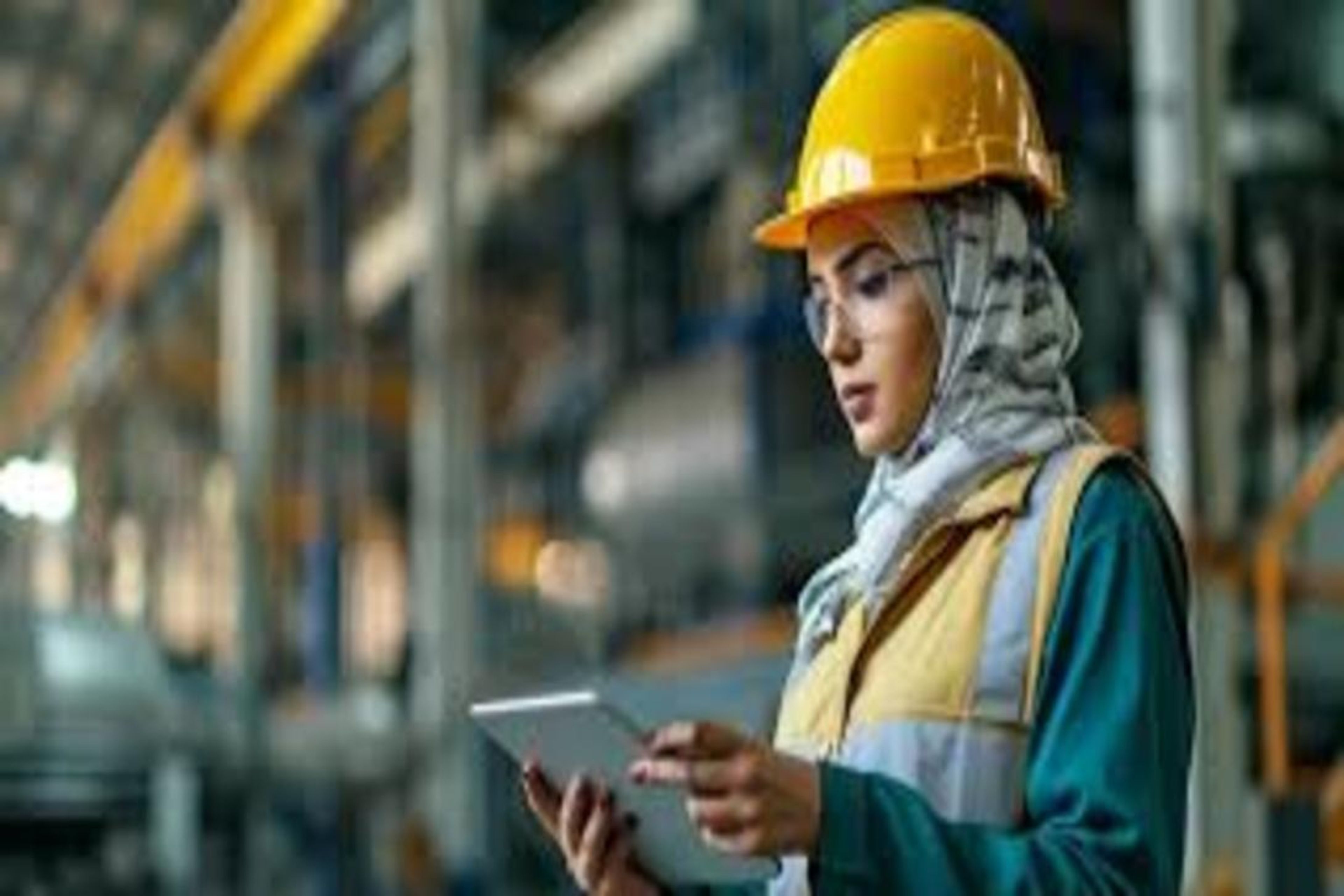 A woman in a yellow hard hat, hijab, and safety vest holds a tablet in an industrial facility.
