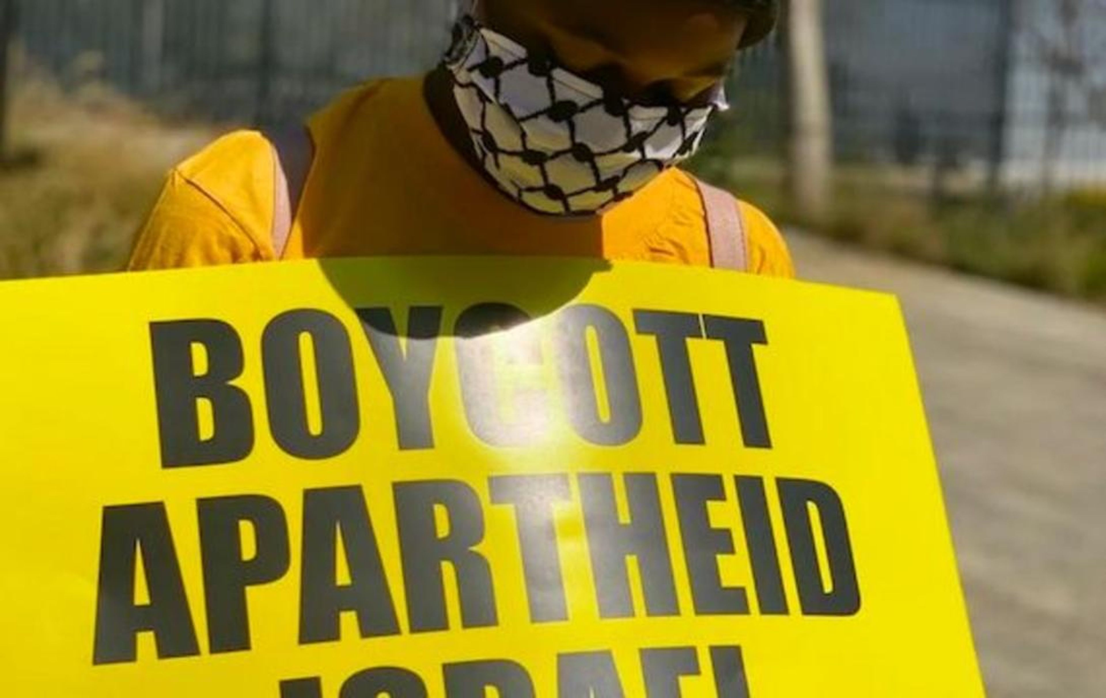 A person wearing a keffiyeh-patterned mask and yellow shirt holds a yellow sign that reads "BOYCOTT APARTHEID ISRAEL."