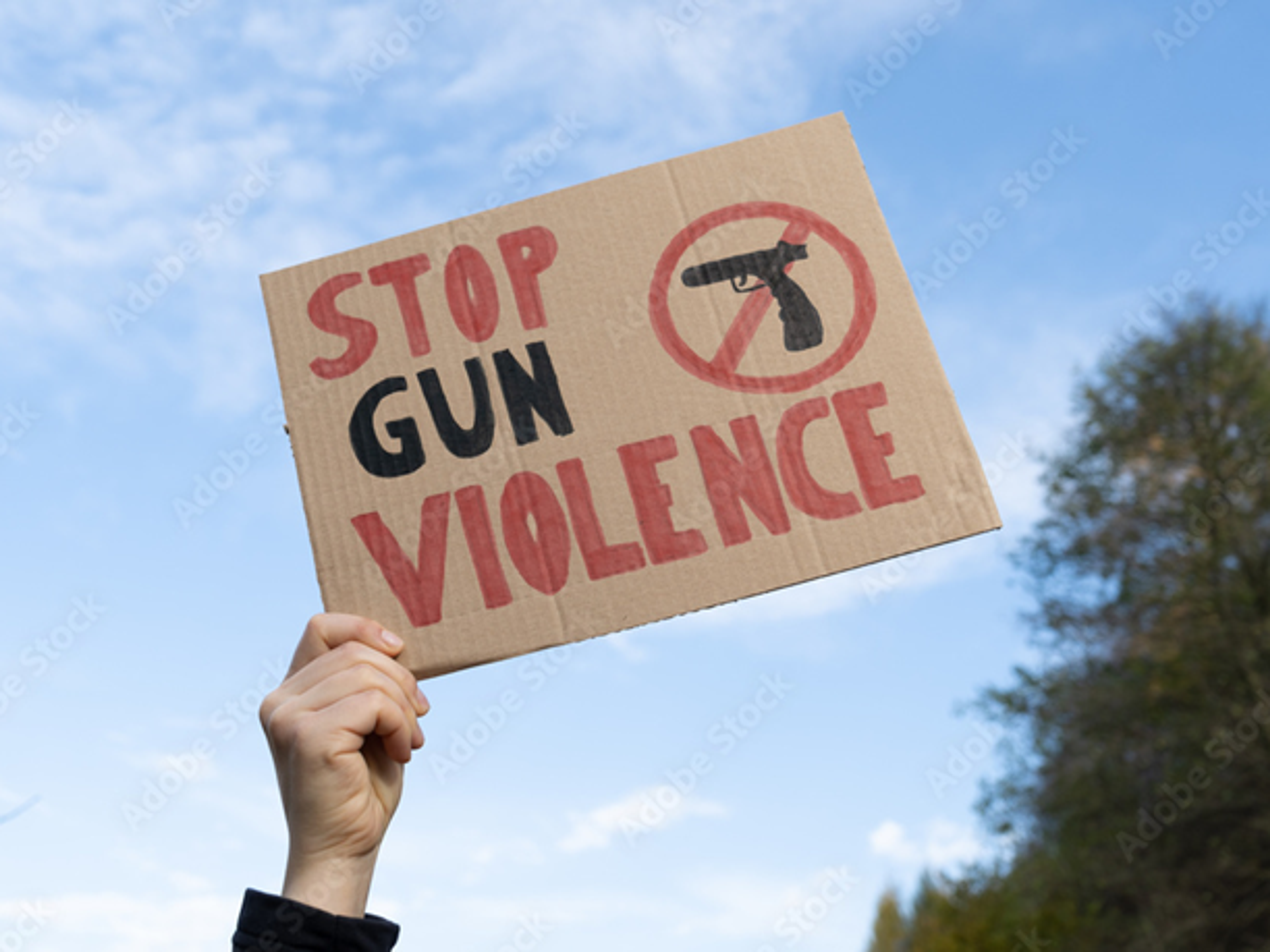A hand holds a cardboard sign that says "STOP GUN VIOLENCE" with a crossed-out gun symbol against a blue sky.