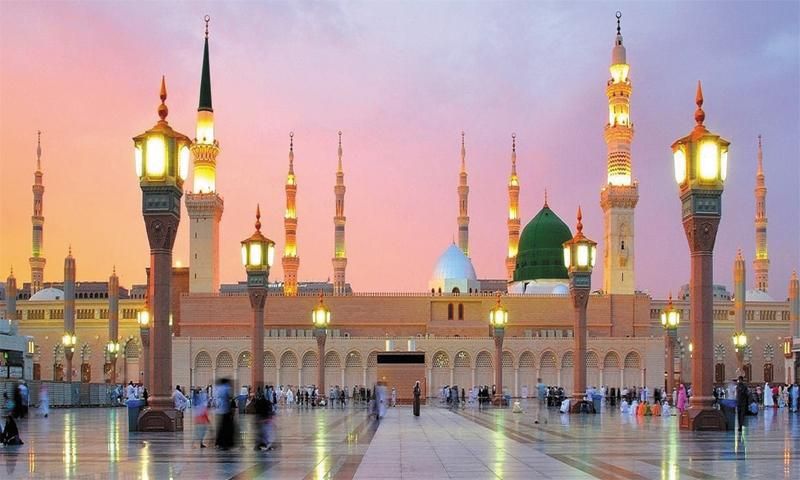 The Prophet's Mosque in Medina at twilight, with its green dome, illuminated minarets, and people in the courtyard.